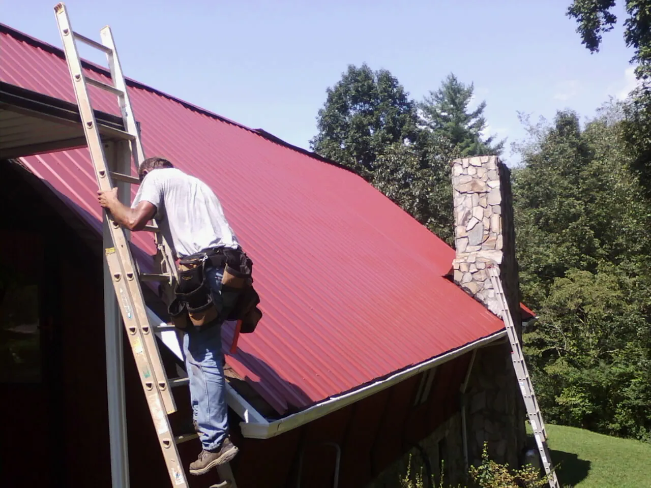 Roofer on a ladder beside a red metal roof and stone chimney — Soffit & Fascia Repair by Roofing Brothers in Middlebrook